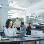 Femme professionnelle tapant sur un ordinateur portable au bureau dans un environnement de bureau à côté d'une imprimante Xerox® C325